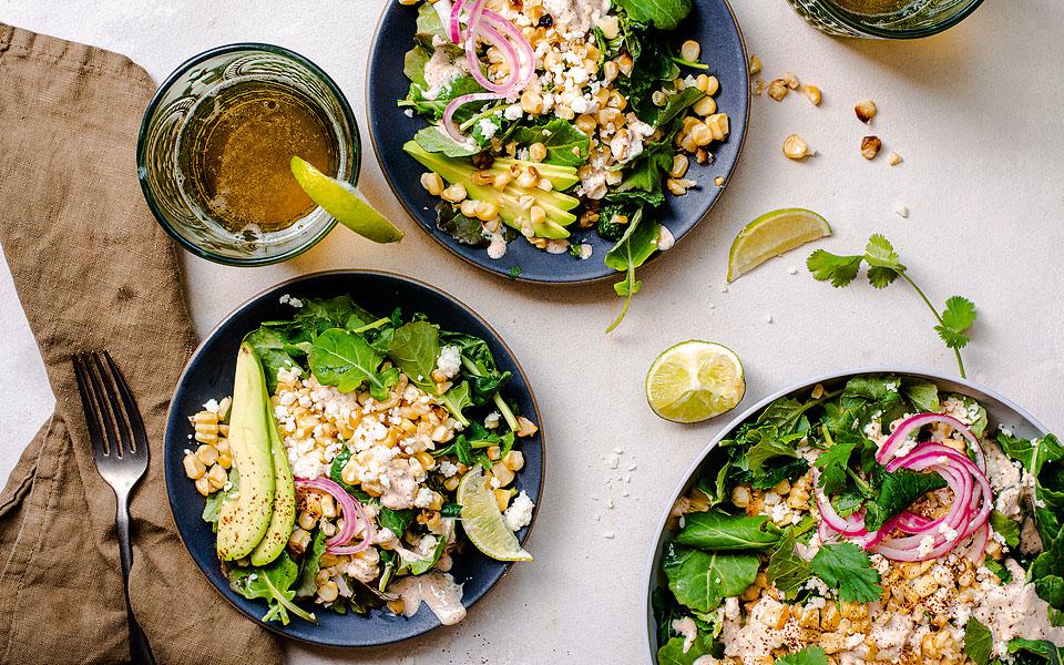 Three bowls of salad with avocado, corn, pickled onions, greens, cheese, and cilantro are on a table with a lime wedge, drink, fork, and brown napkin nearby.