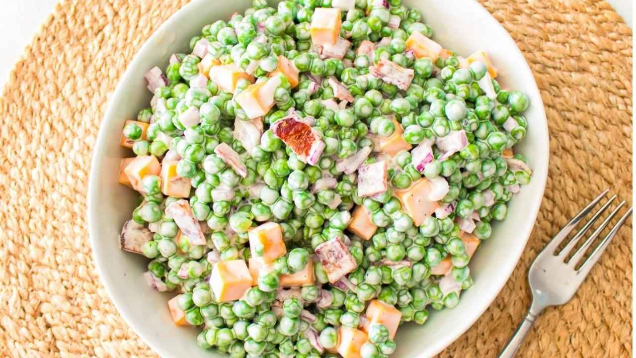A bowl of pea salad with diced cheddar cheese, bacon bits, red onion, and creamy dressing, placed on a woven placemat with a fork beside it.