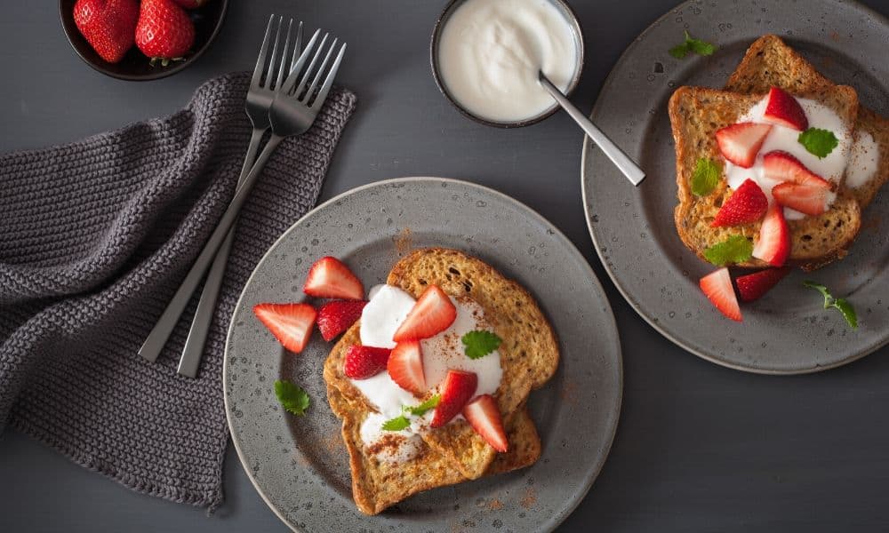Two plates of French toast topped with whipped cream, sliced strawberries, and mint leaves, next to a bowl of strawberries, a bowl of yogurt, utensils, and a gray napkin.
