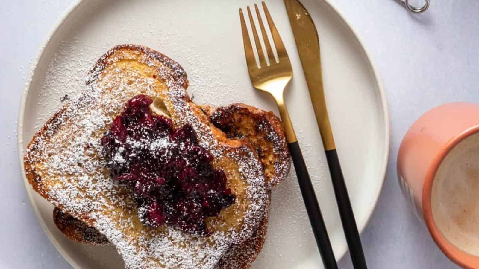 Two slices of French toast topped with berry compote and powdered sugar on a white plate, with a fork and knife. A cup of coffee is next to the plate.
