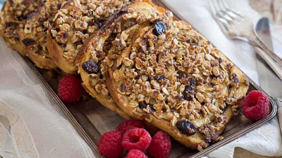 Sliced baked French toast topped with granola and dried fruit, served on a tray with fresh raspberries, next to forks and a white napkin.