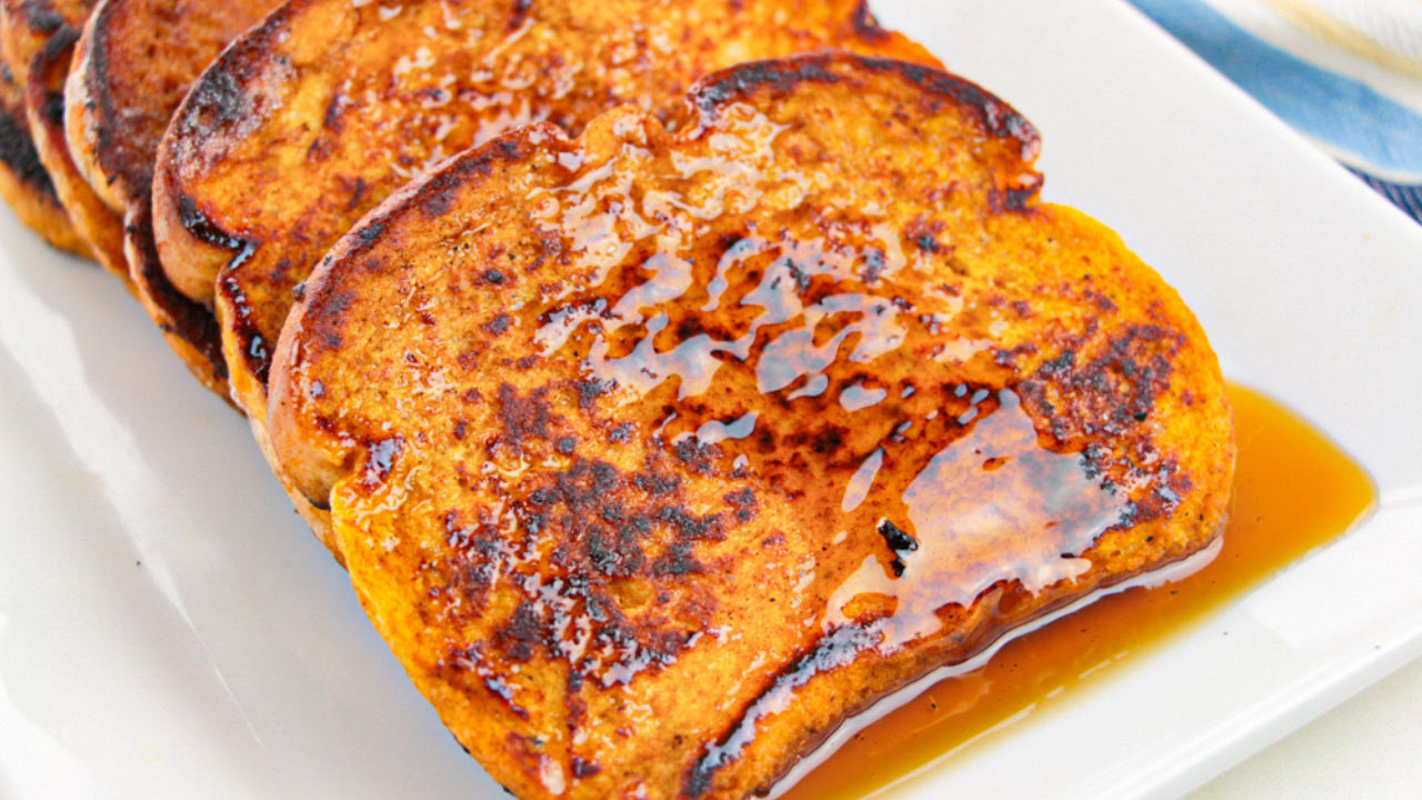 Four slices of French toast with a golden-brown crust are arranged on a white plate and topped with syrup.