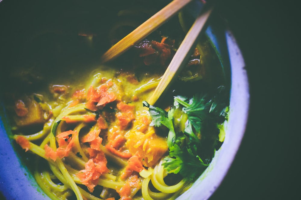 A bowl of noodle soup with chopped herbs and vegetables, topped with grated ingredients, and two wooden chopsticks resting inside.
