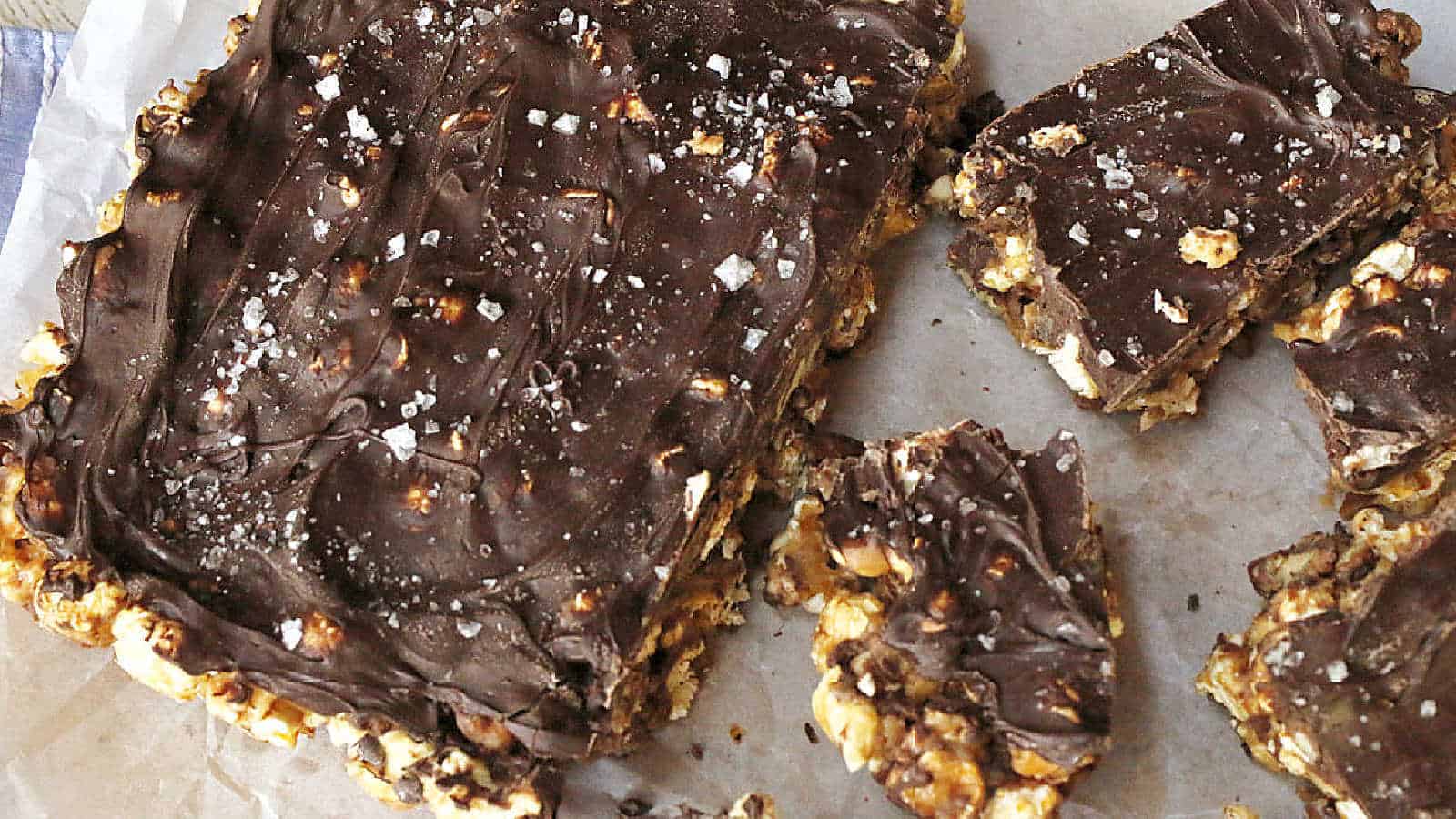 A large rectangular chocolate-topped dessert bar with a popcorn and nut base, cut into smaller pieces, sprinkled with coarse salt, on a sheet of parchment paper.