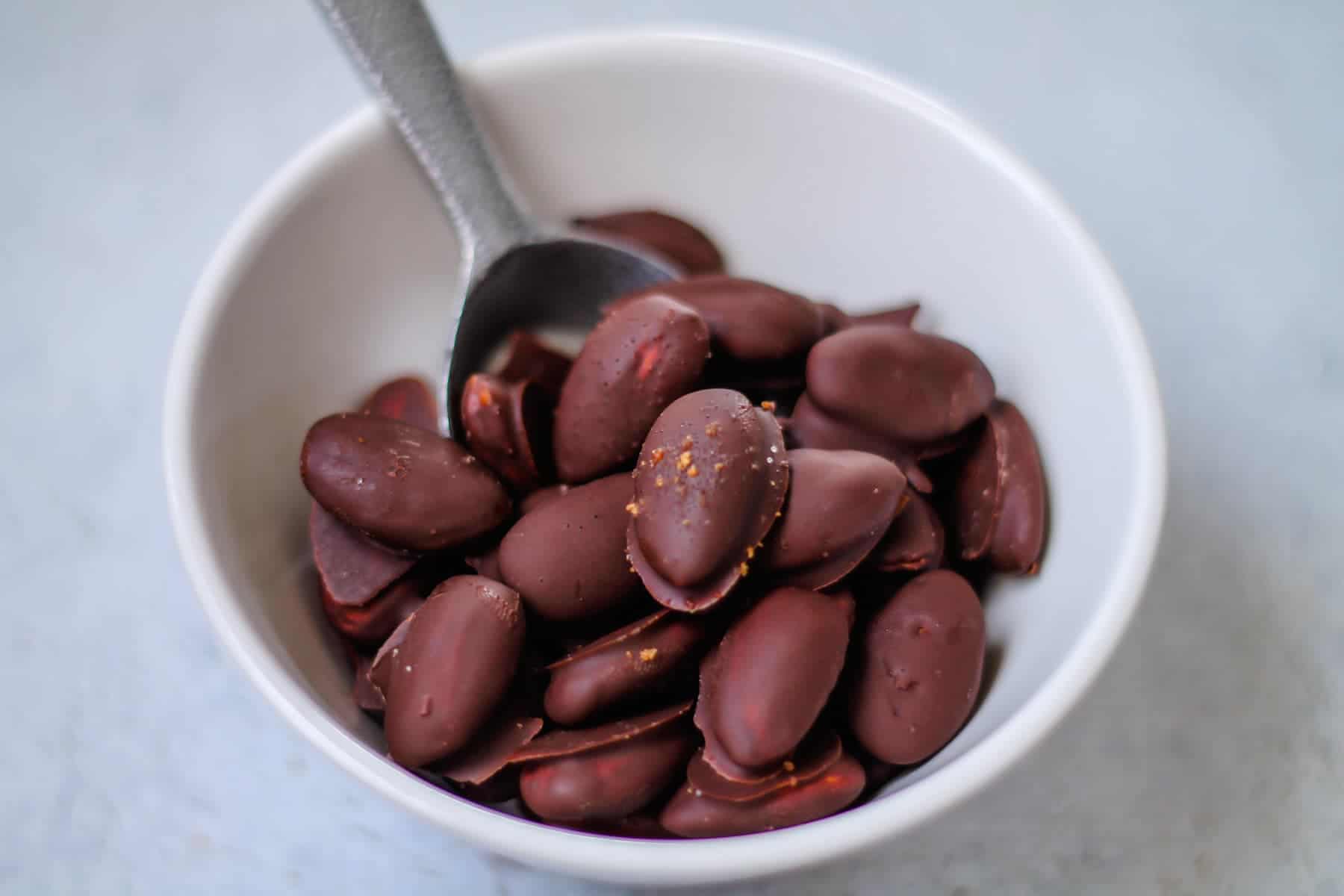 A white bowl filled with chocolate-covered almonds with a metal spoon resting inside.