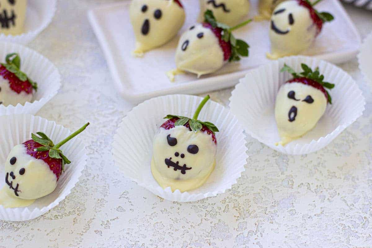 Strawberries dipped in white chocolate with faces drawn in dark chocolate, arranged in paper cups and on a white platter.