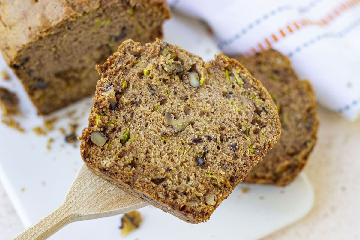 A close-up of a slice of zucchini bread with nuts being lifted by a wooden spatula, with more bread slices in the background.