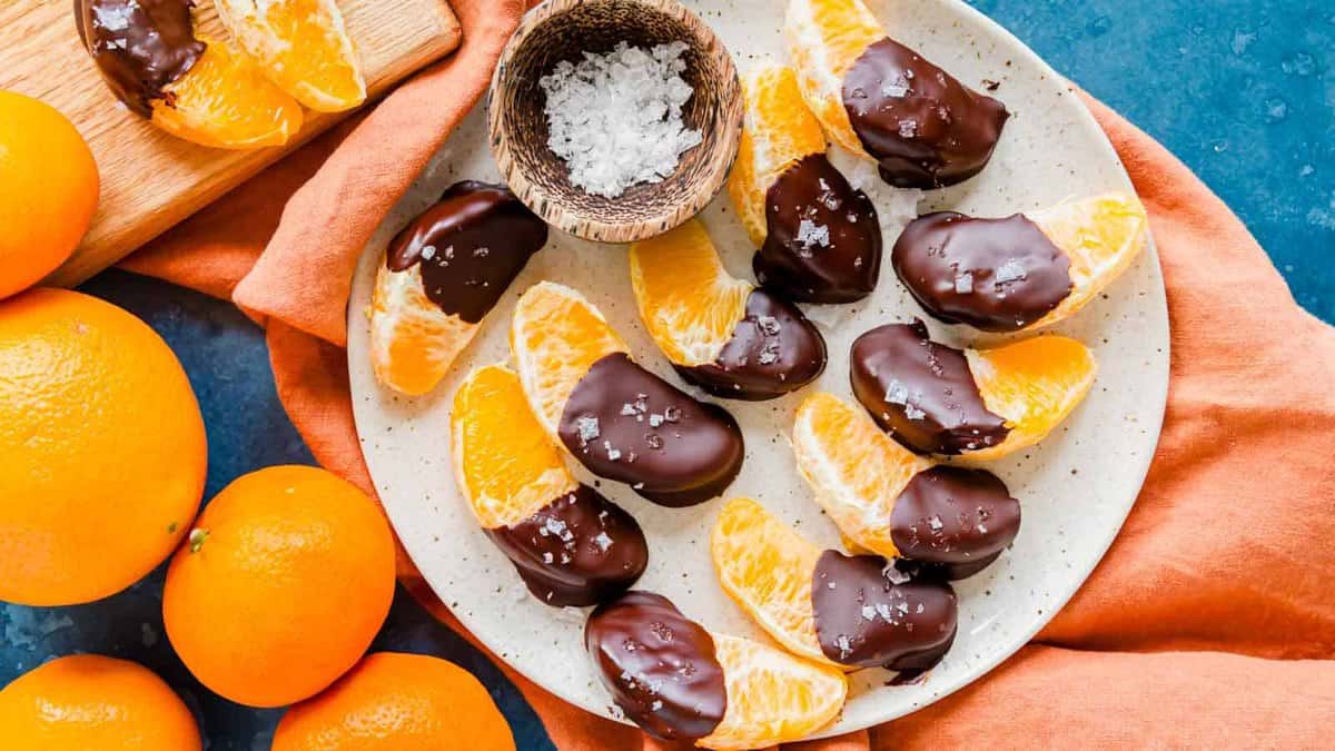 Orange segments dipped in chocolate and sprinkled with sea salt are arranged on a speckled plate, with whole oranges and an orange napkin nearby.