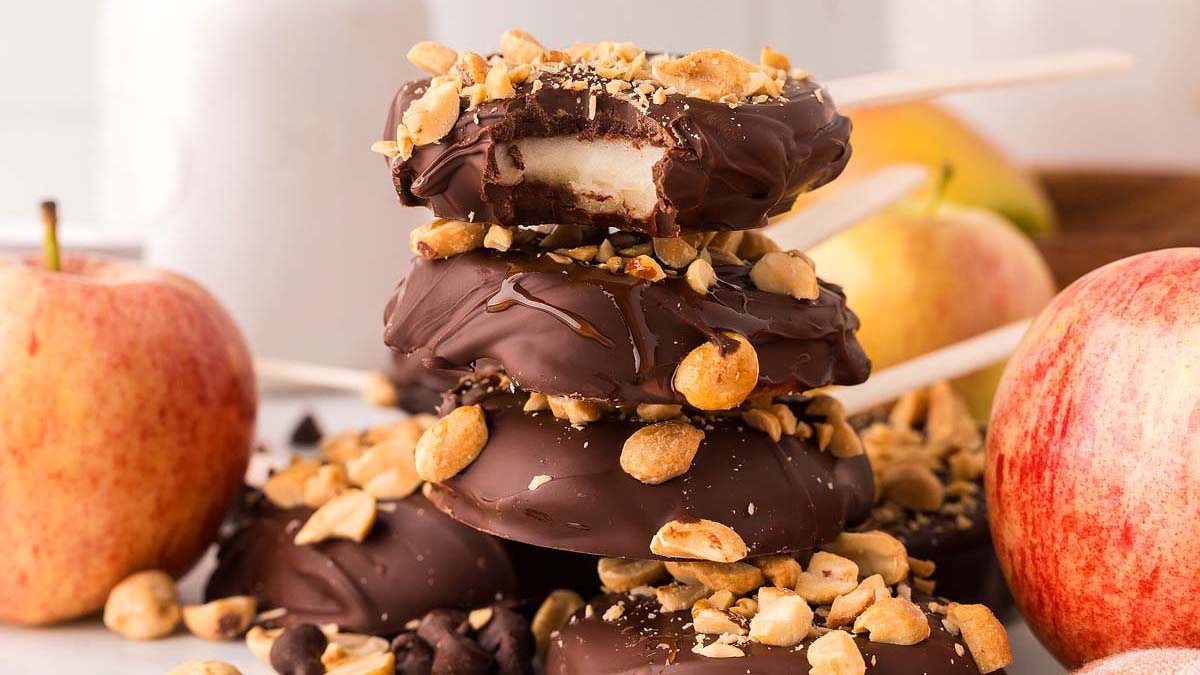 Stack of chocolate-covered apple slices on sticks, topped with chopped peanuts, surrounded by whole apples and more nuts.
