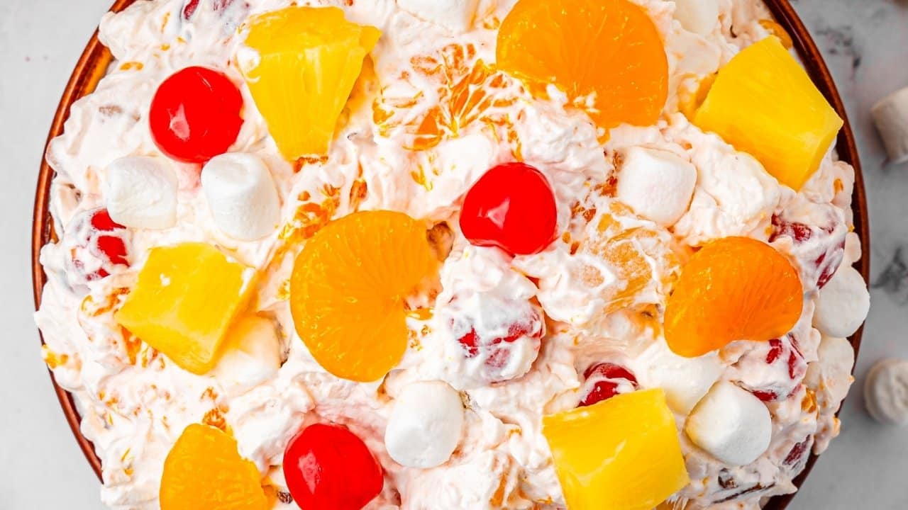 A bowl of fruit salad with whipped cream, mini marshmallows, maraschino cherries, pineapple chunks, and mandarin orange segments.