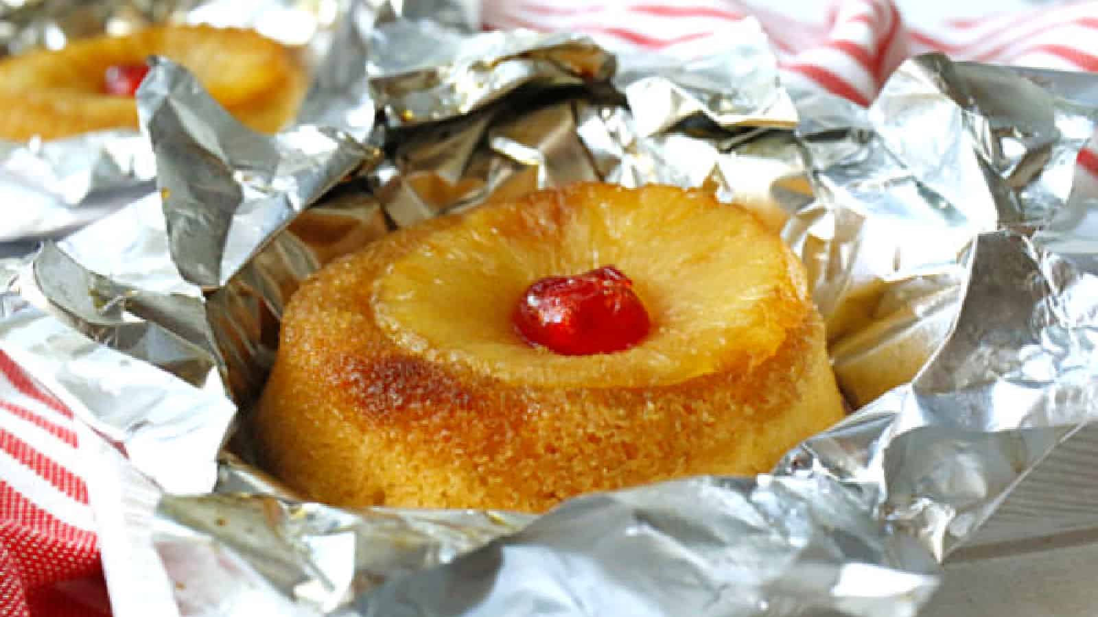 A pineapple upside-down cake with a cherry on top sits partially unwrapped in aluminum foil.