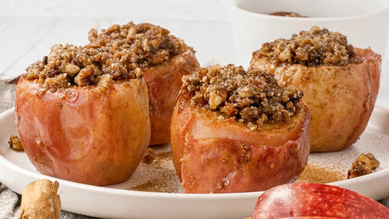 Three baked apples filled with a crumbly nut and spice mixture are arranged on a white plate, with a small bowl in the background.