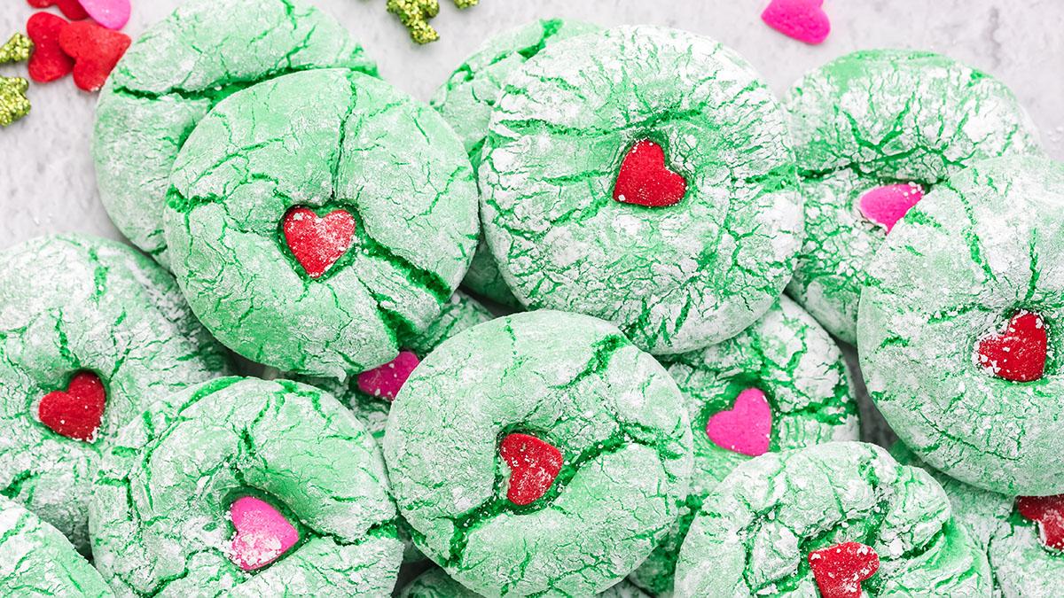 Green crinkle cookies dusted with powdered sugar, each topped with a red or pink heart-shaped candy, arranged closely together on a light surface.