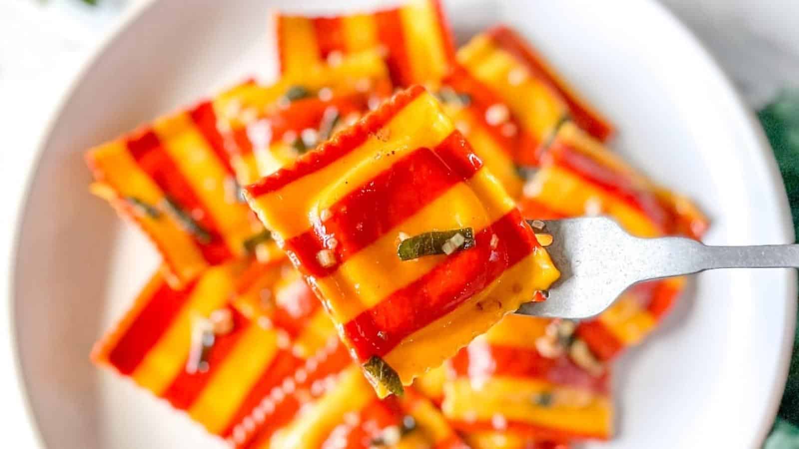 A close-up of striped red and yellow ravioli garnished with herbs, served on a white plate with one piece held up by a fork.