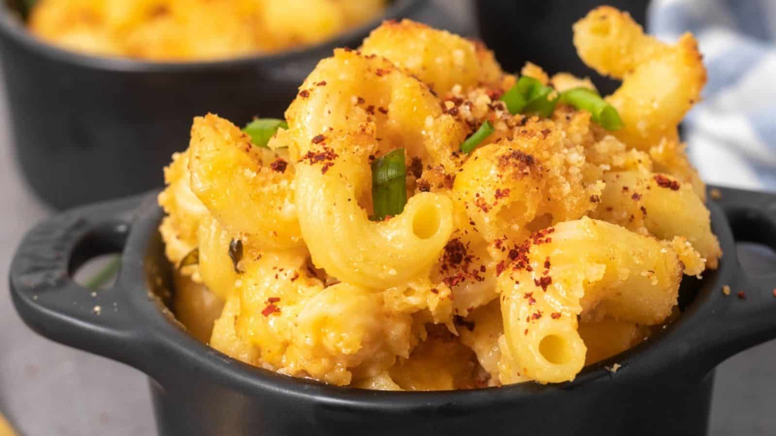A black bowl filled with baked macaroni and cheese, topped with breadcrumbs, chopped chives, and red pepper flakes.