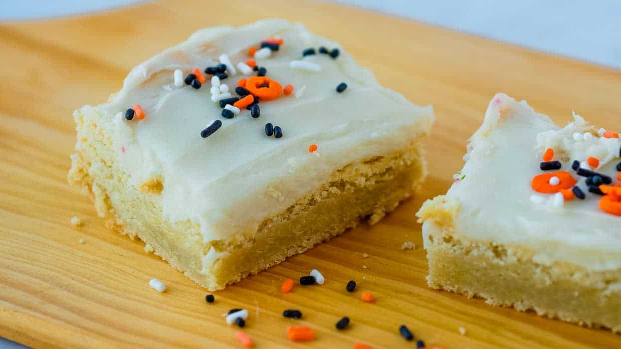 A close-up of two frosted sugar cookie bars with white icing and black, white, and orange sprinkles on a wooden board.