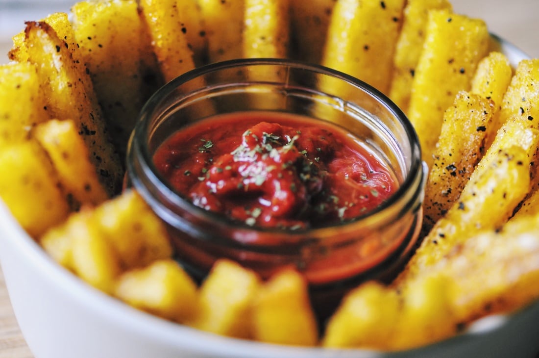 A bowl of seasoned fries arranged around a small glass container of tomato dipping sauce sprinkled with herbs.