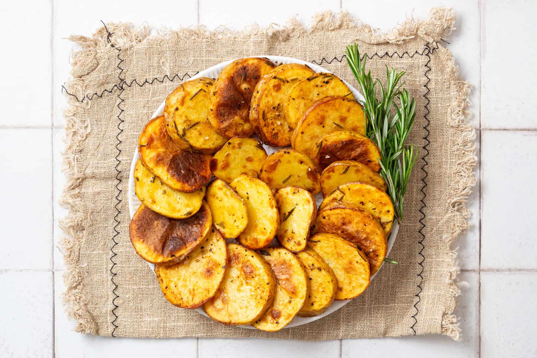 A white plate with roasted, seasoned potato slices arranged in a circle, garnished with fresh rosemary sprigs, on a piece of burlap fabric.