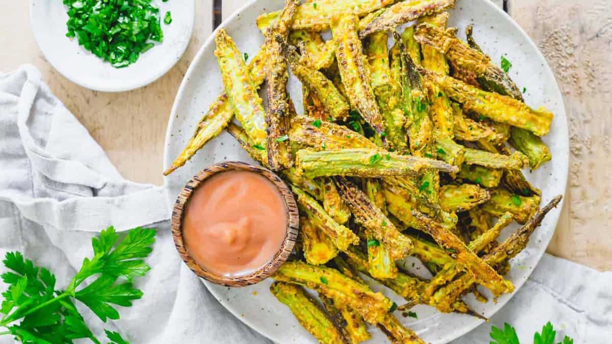 A plate of crispy baked okra fries with chopped herbs, served with a small bowl of dipping sauce on the side.