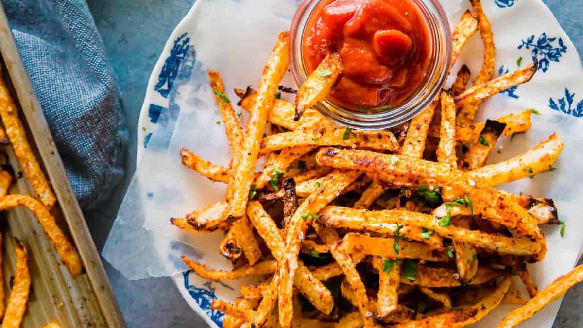 A plate of baked fries garnished with herbs, served with a small glass jar of ketchup on the side.