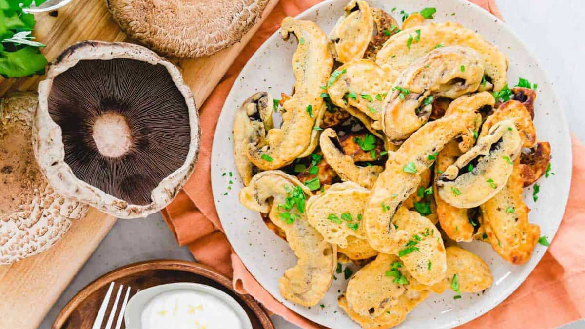 A plate of golden-brown fried mushroom slices garnished with chopped herbs, next to whole portobello mushrooms and a small bowl of white dipping sauce.