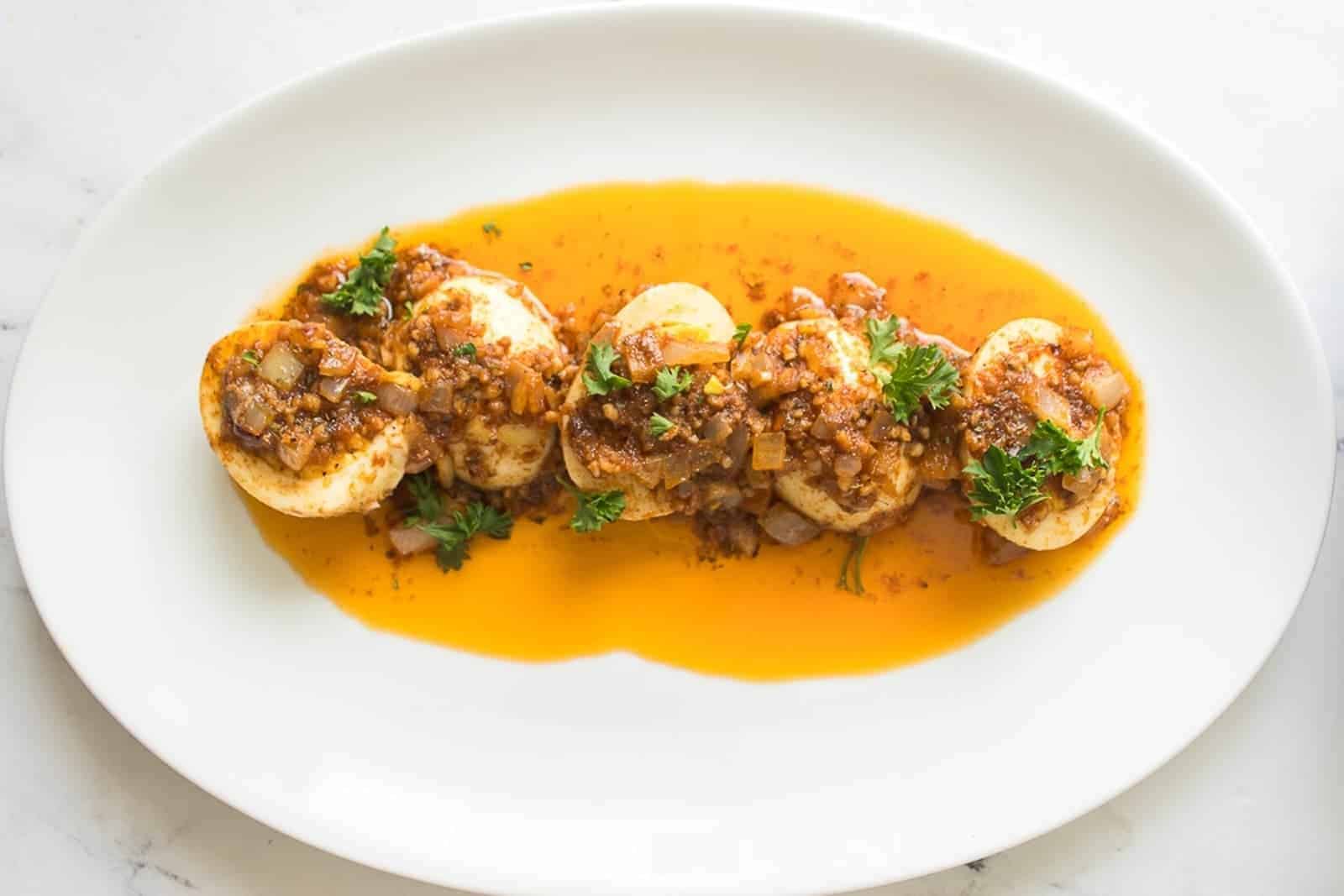 Four halved boiled eggs topped with a chunky, reddish-brown sauce and garnished with parsley, served on a white oval plate.