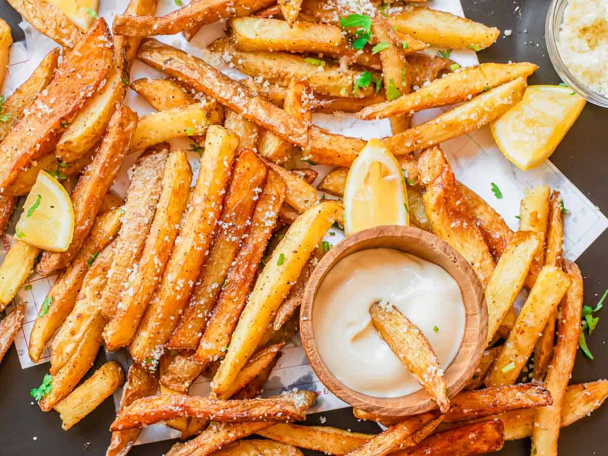 A pile of golden-brown French fries garnished with parsley and grated cheese, served with lemon wedges and a bowl of dipping sauce.