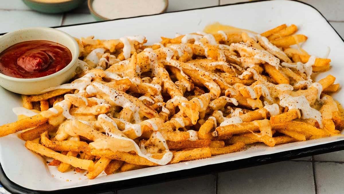 A tray of seasoned French fries topped with cheese sauce and white drizzle, served with a side of ketchup.