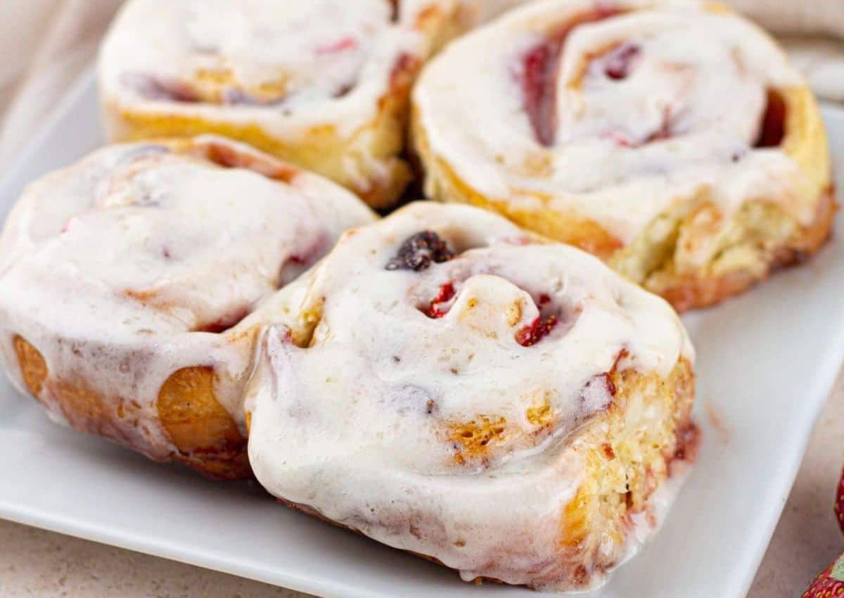 Four frosted cinnamon rolls with swirled filling are arranged on a white square plate.