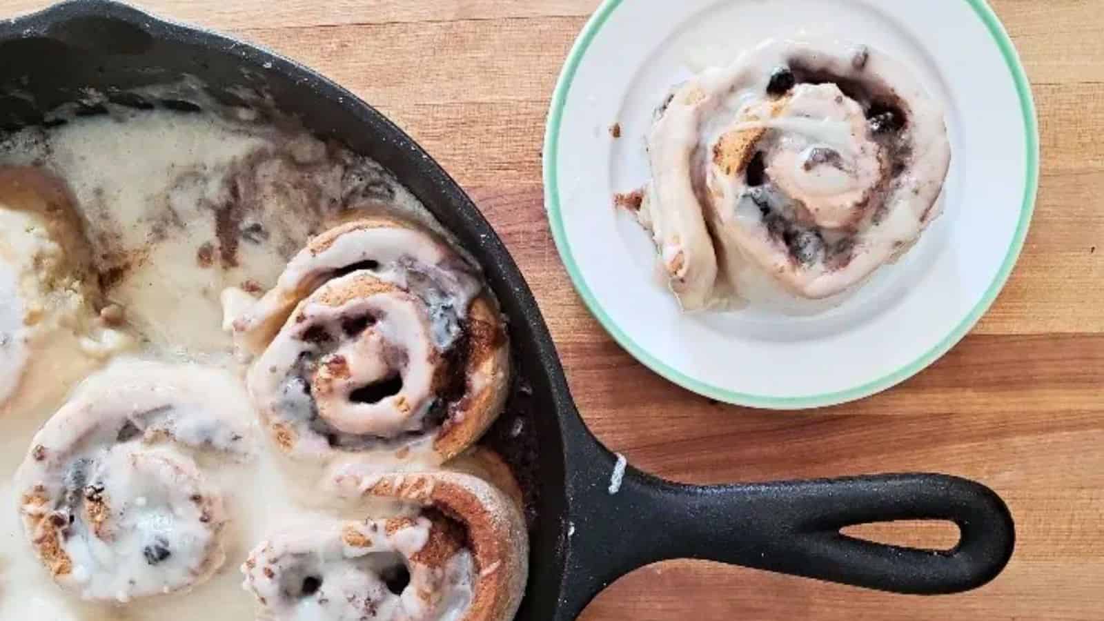 A skillet with frosted cinnamon rolls, with one cinnamon roll served on a small white plate nearby, on a wooden surface.