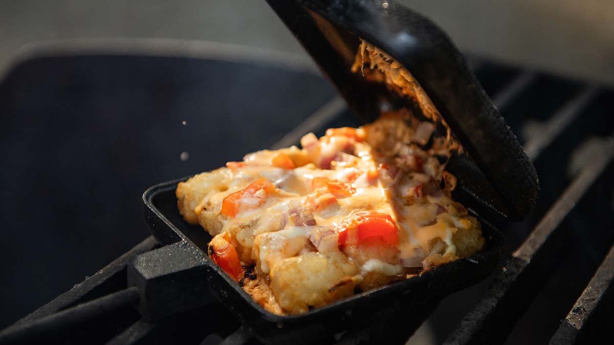 A close-up of a pie iron being opened over a grill, revealing melted cheese, diced tomatoes, and tater tots inside.