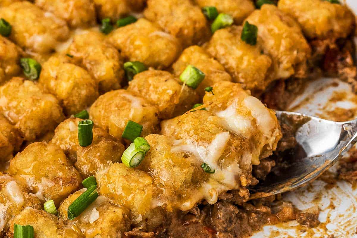A serving of tater tot casserole with melted cheese and ground meat, topped with chopped green onions, being lifted from a baking dish.