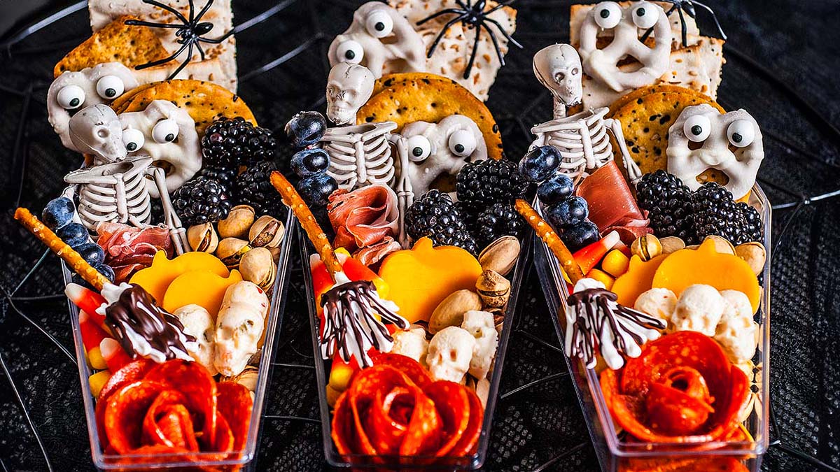 Three Halloween-themed snack cups with crackers, cheese, meats, berries, nuts, and skeleton decorations, arranged on a dark surface with fake spiders.