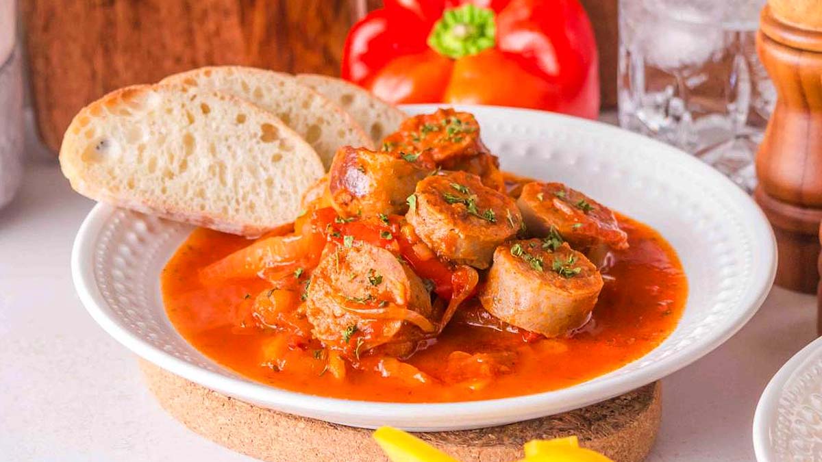 A white plate with sliced sausage in tomato sauce, garnished with herbs, served with two slices of bread. A red bell pepper and glass of water are in the background.