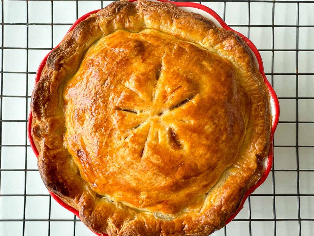 A golden-brown baked pie with a flaky crust and decorative slits, cooling on a black wire rack.