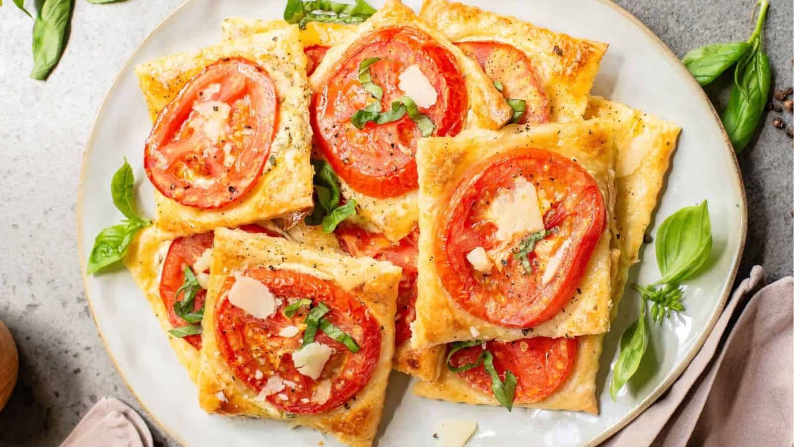 A plate of rectangular puff pastry tarts topped with sliced tomatoes, basil, and shaved cheese sits on a table with fresh basil leaves scattered around.