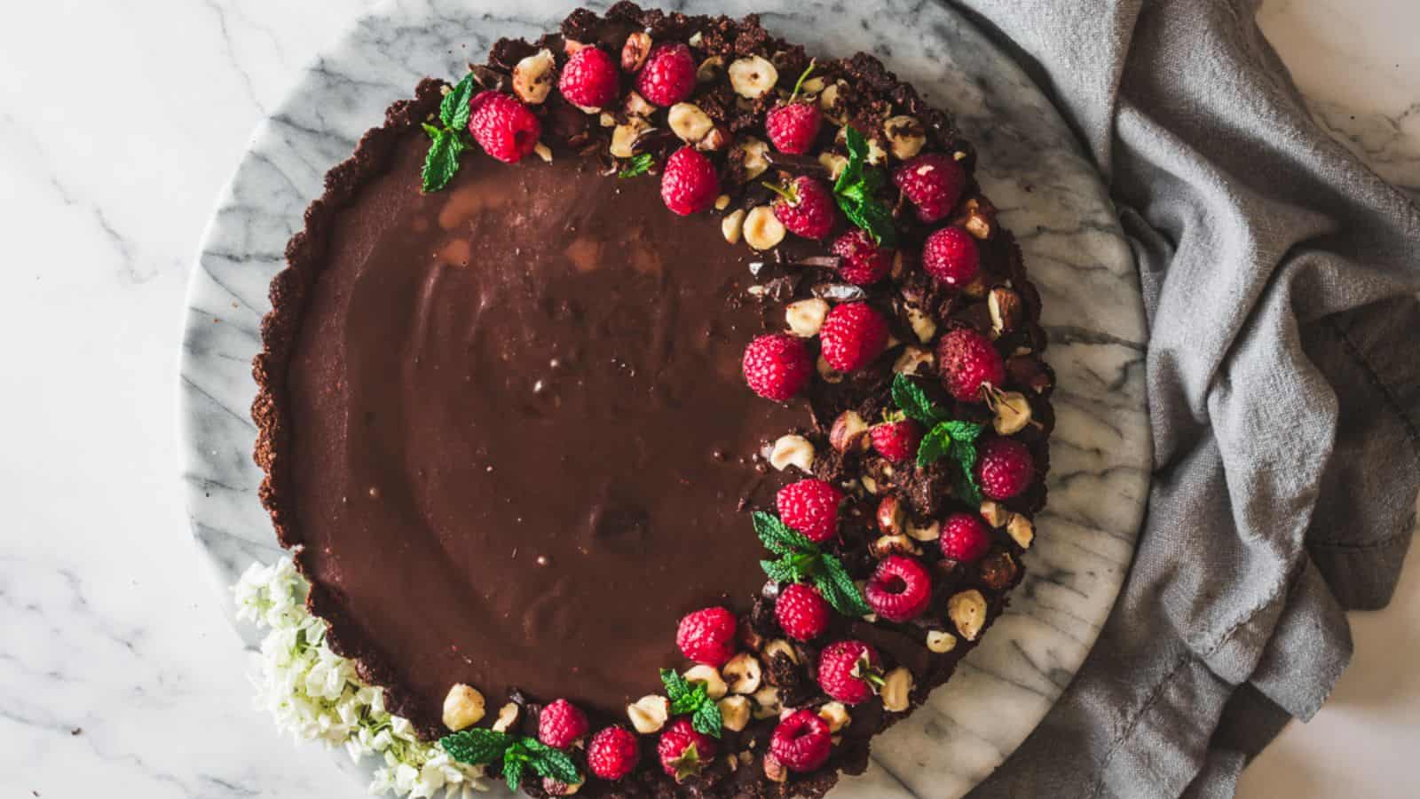 A chocolate tart on a marble surface, half decorated with raspberries, hazelnuts, mint leaves, and a cluster of white flowers, next to a gray cloth.