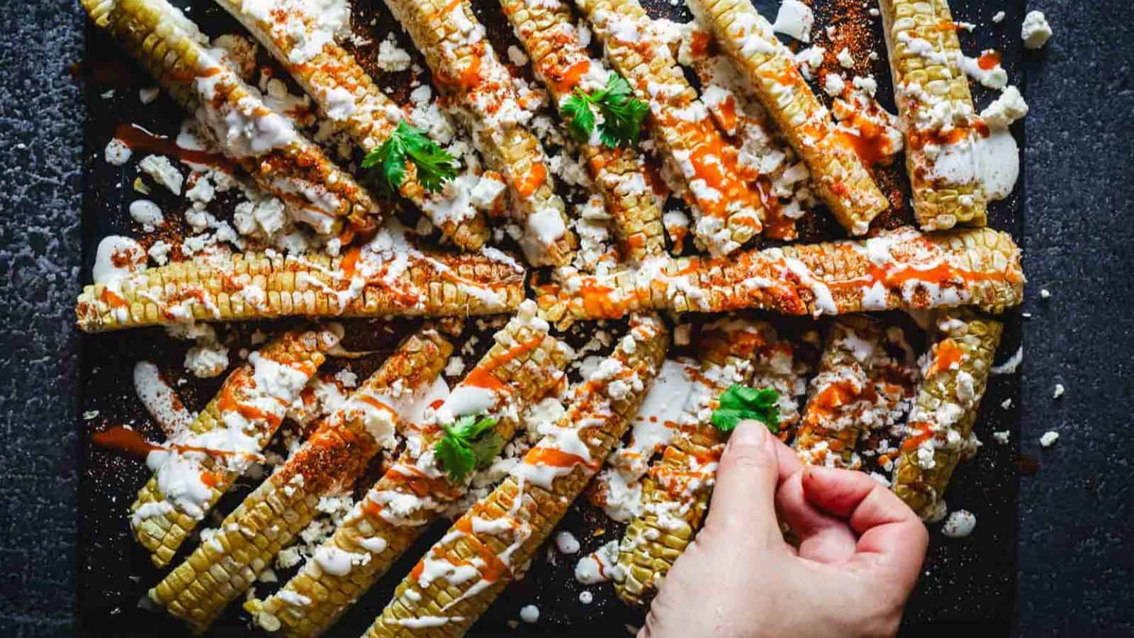 Rows of grilled corn ribs topped with white sauce, cheese, chili powder, and herbs are arranged on a dark surface. A hand is placing a sprig of fresh cilantro on top.