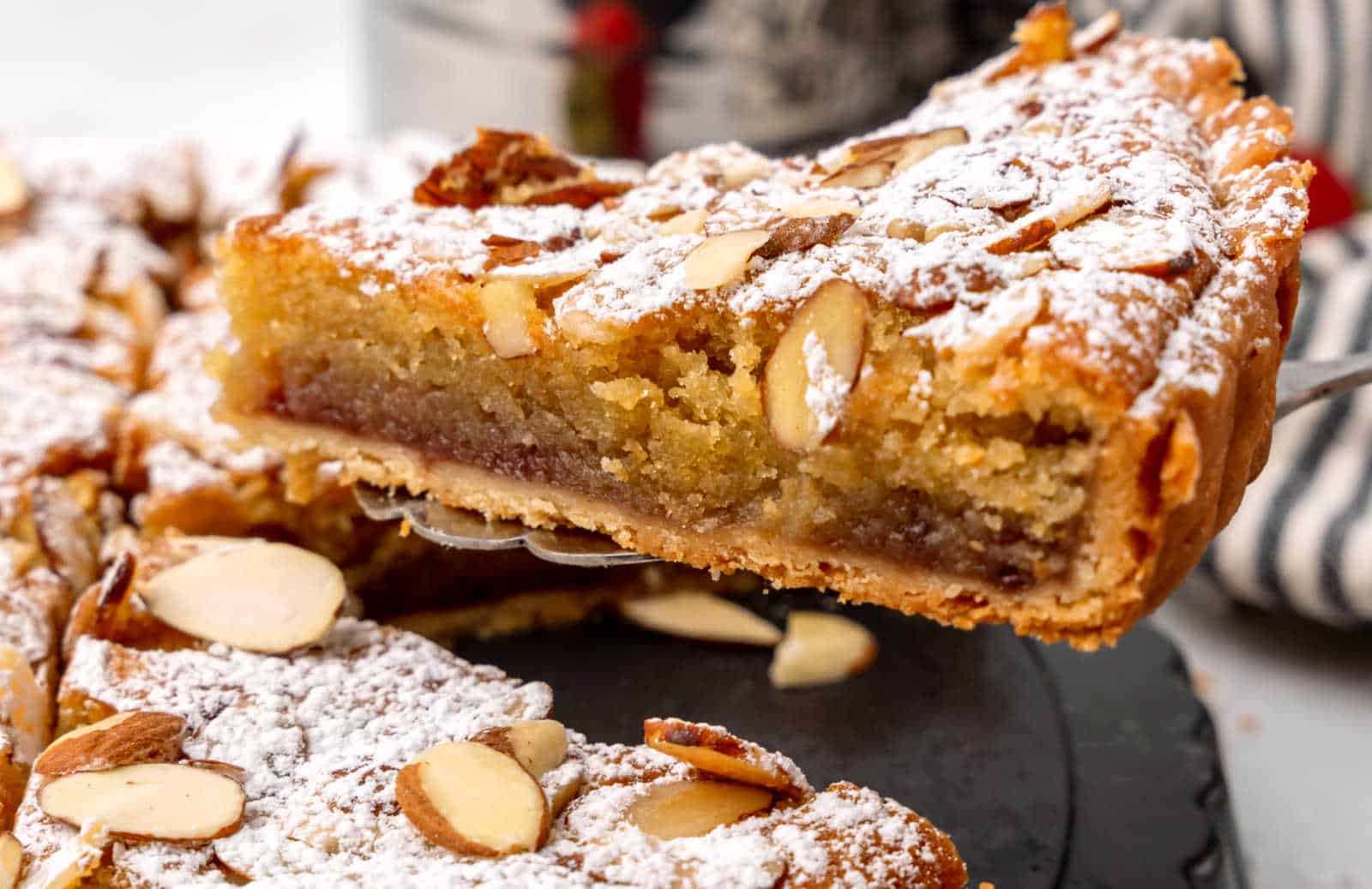 A close-up of a slice of almond tart topped with sliced almonds and powdered sugar, showing a golden, moist interior.