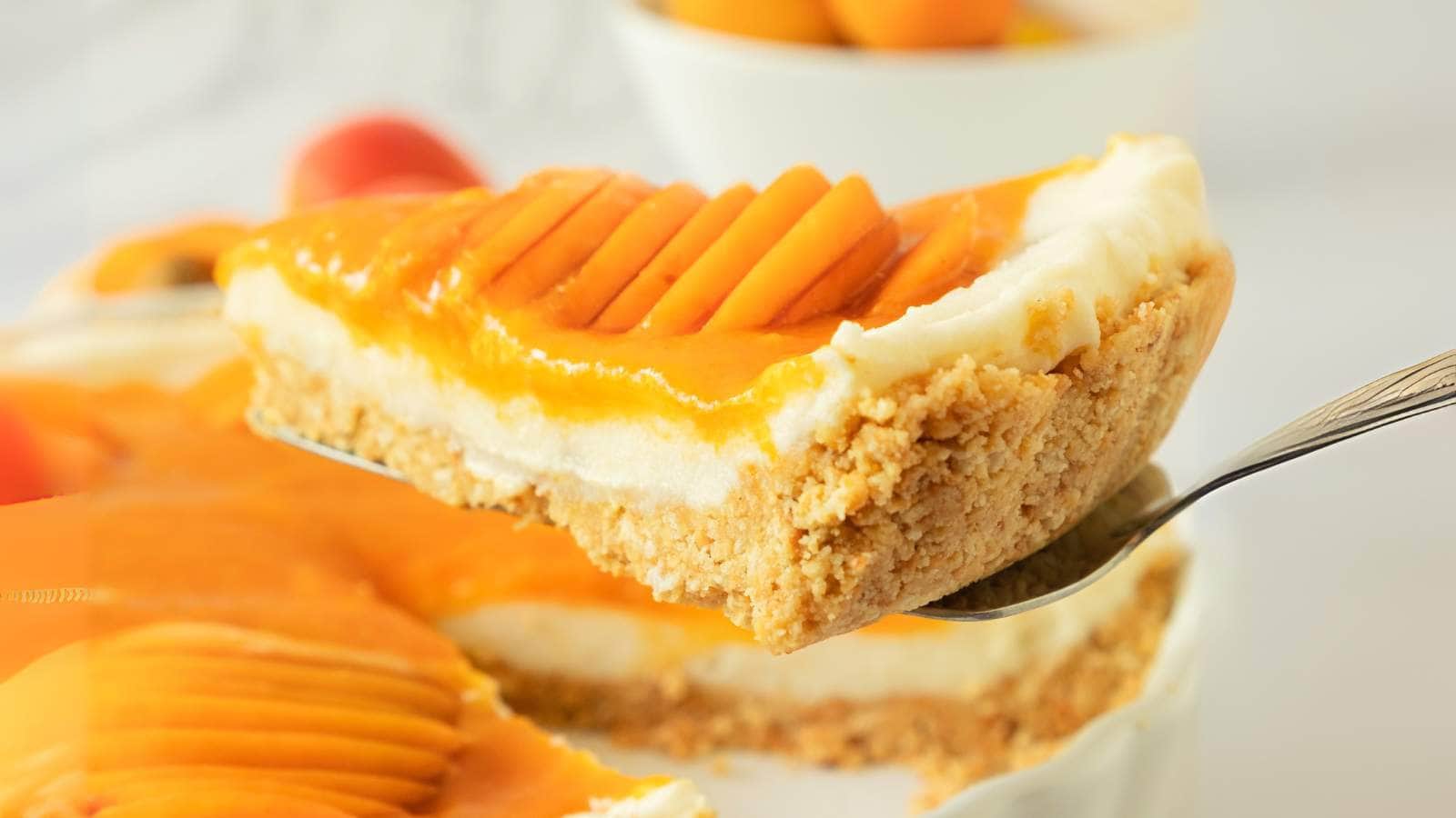 A slice of mango cheesecake with a crumbly crust and creamy filling, topped with sliced fresh mango, held up on a serving spatula.