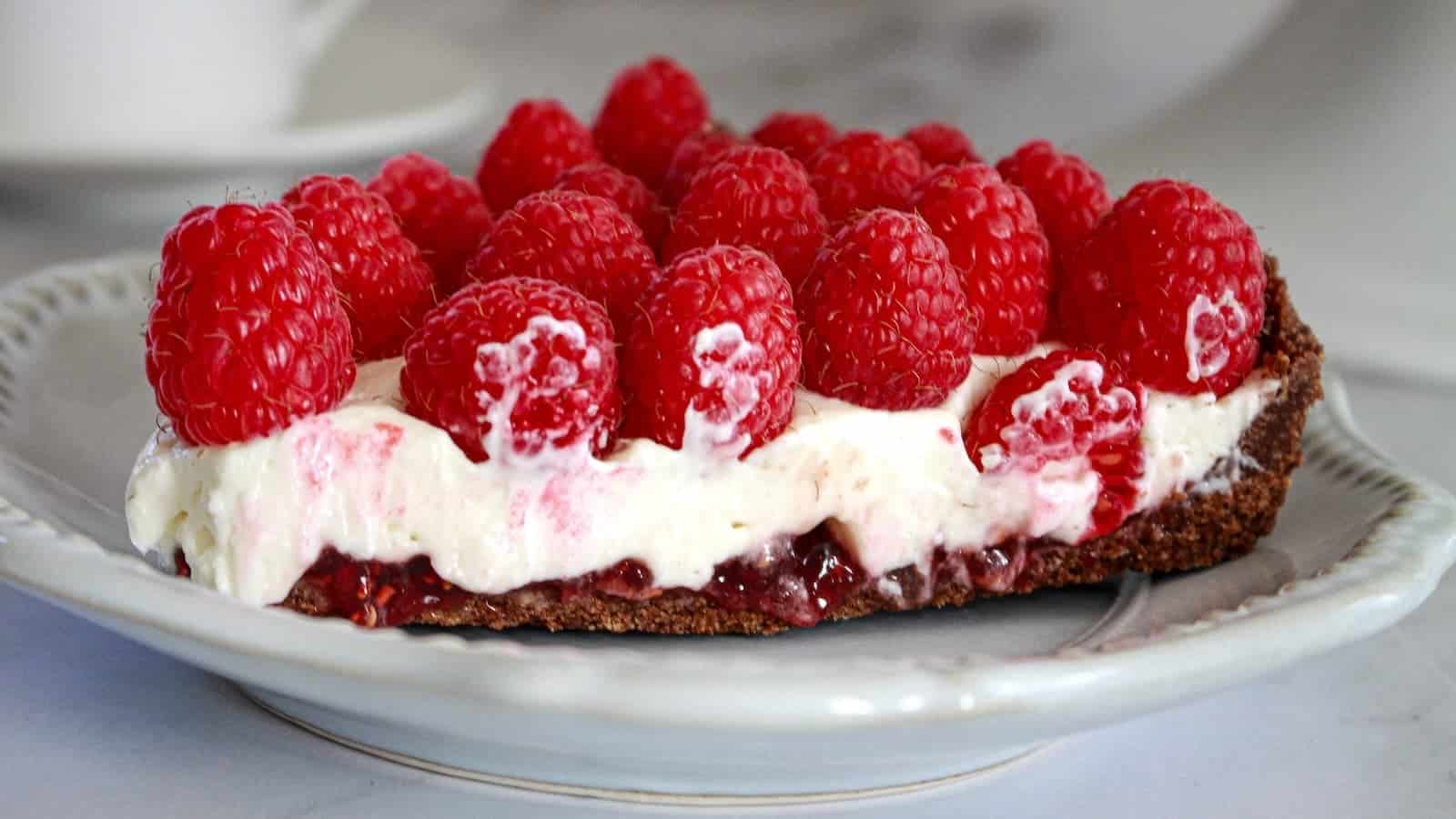 A slice of dessert tart with a chocolate crust, creamy filling, raspberry jam layer, and topped with fresh raspberries on a white plate.