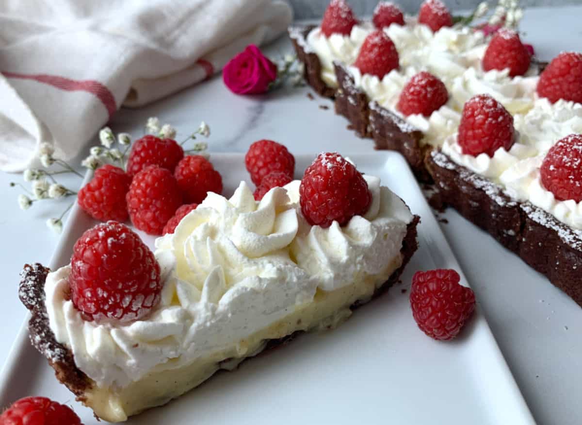 A slice of tart with whipped cream and fresh raspberries on a plate, with more raspberries and a whole tart in the background.