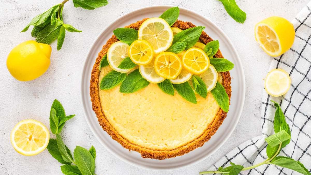 A lemon tart garnished with fresh mint leaves and lemon slices on a white plate, surrounded by lemons and mint on a white surface.