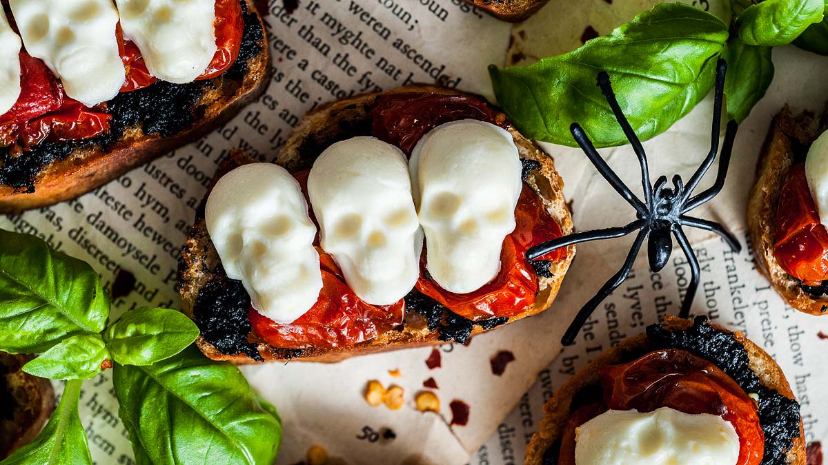 Toasted bread topped with roasted tomatoes and skull-shaped mozzarella, surrounded by basil leaves and a plastic spider on a newspaper background.