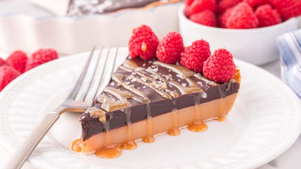 A slice of chocolate and caramel tart topped with raspberries and drizzled with caramel sauce sits on a white plate with a fork.