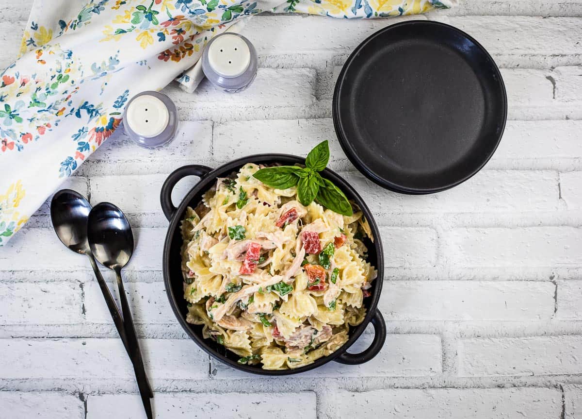 A black bowl filled with bowtie pasta salad, garnished with basil, sits on a white brick surface next to a black plate, two spoons, and salt and pepper shakers.