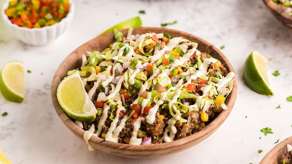 A wooden bowl filled with a quinoa salad topped with diced vegetables, avocado, and a drizzle of creamy dressing, with lime wedges on the side.