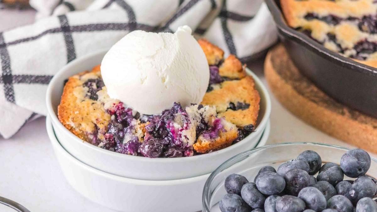 A bowl of blueberry cobbler topped with a scoop of vanilla ice cream, with fresh blueberries and a checkered towel in the background.