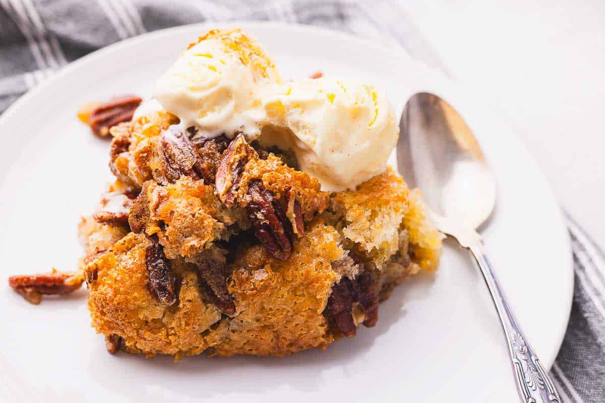 A serving of pecan cobbler topped with two scoops of vanilla ice cream on a white plate with a silver spoon beside it.