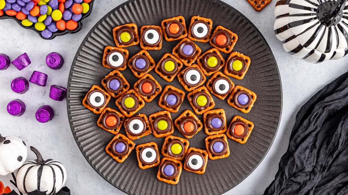 A plate of pretzel bites topped with colorful candies and candy eyes, surrounded by Halloween-themed decorations and treats.